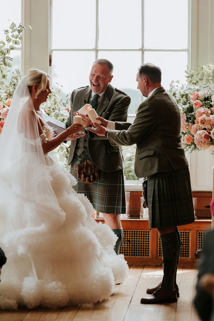 bride in large fluffy wedding dress, groom and celebrant in kilts, all three hold out candles to light at wedding ceremony 