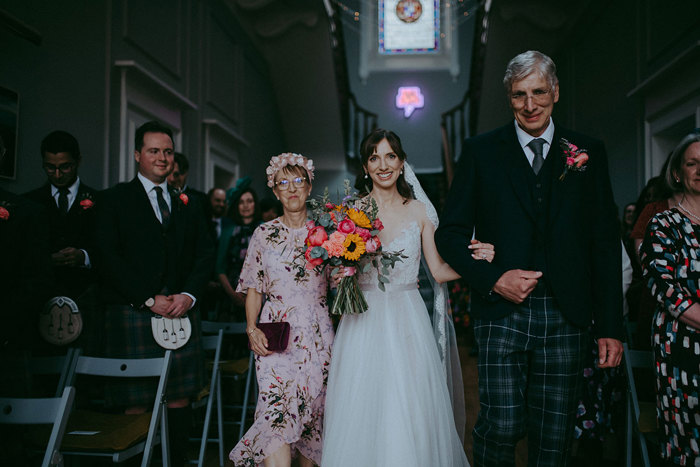 a bride walking down the aisle in between a man in a kilt and woman wearing a pink floral dress