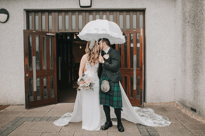 Bride and groom kiss under a white umbrella outside church