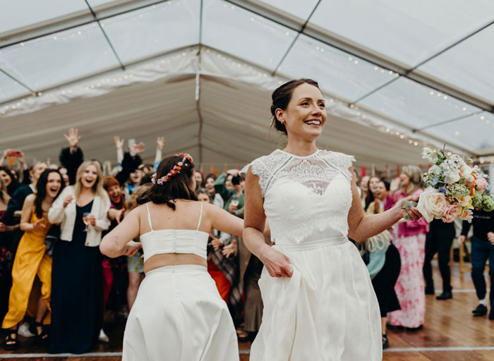 a bride prepares to throw a bouquet with second bride and surprised guests behind her looking animated, some with hands outstretched trying to catch it
