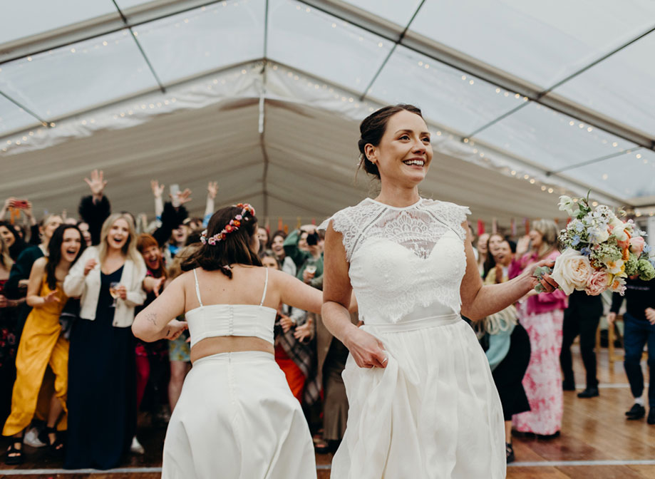 a bride prepares to throw a bouquet with second bride and surprised guests behind her looking animated, some with hands outstretched trying to catch it
