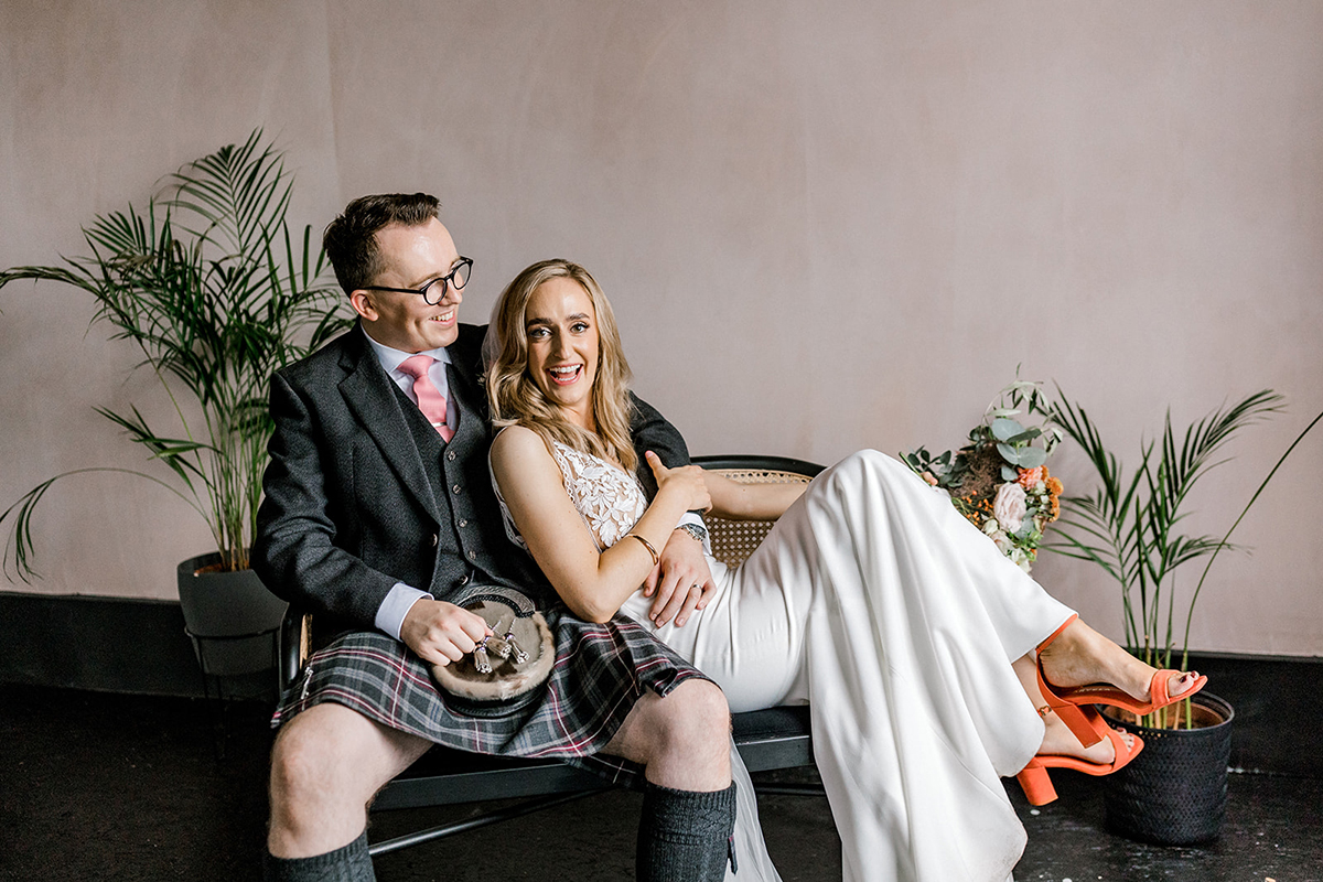 Bride in white wedding outfit and groom in tartan kilt sit together on a bench with potted plants and a blush wall behind them.