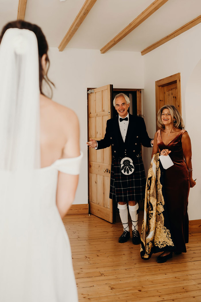 a man in a kilt and a woman in a velvet dress standing in a doorway looking at a bride