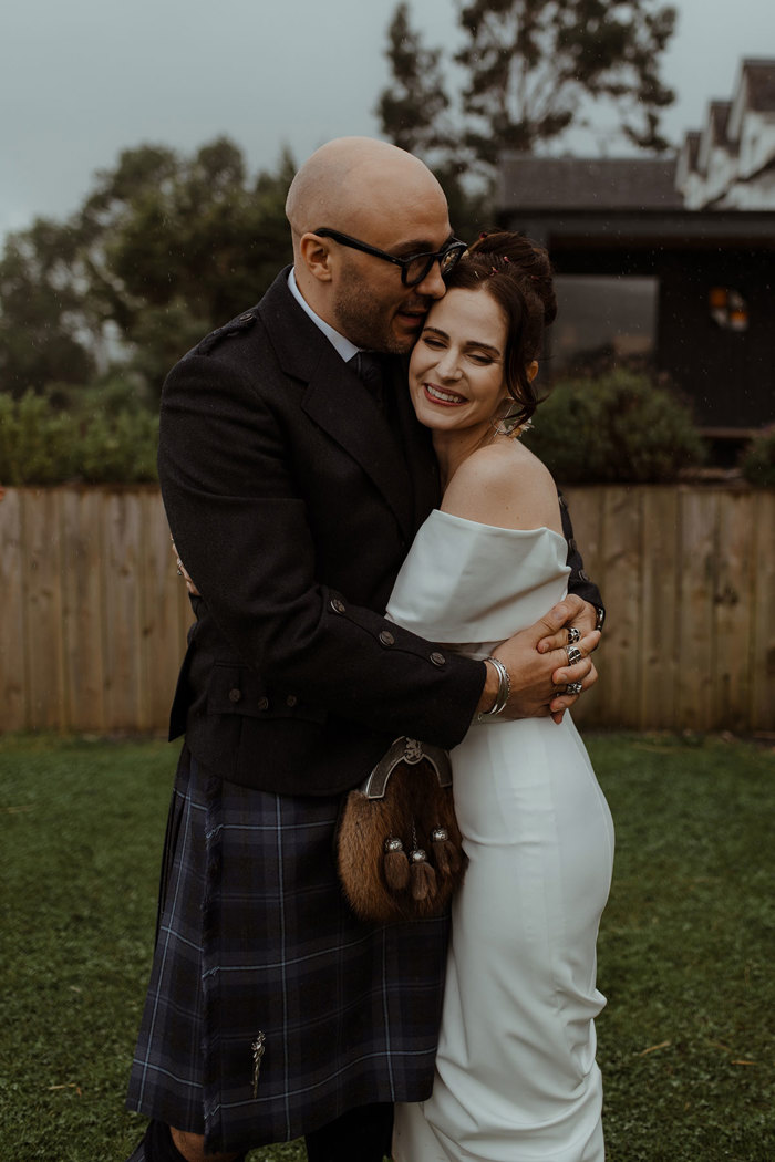 groom in blue, grey and white tartan kilt outfit stands outdoors, wrapping his arms around waist of smiling bride in off the shoulder white wedding dress