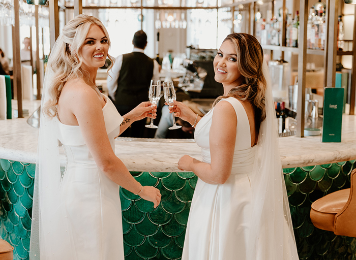 two brides hold champagne glasses standing against a bar at Kimpton Blythswood Square Hotel