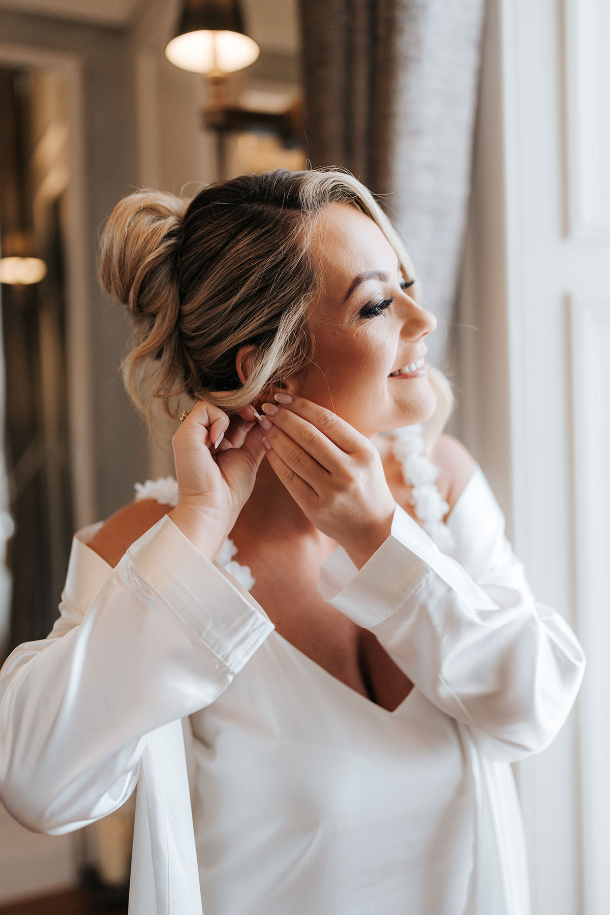 Bride smiling as she puts on her earrings by the window during wedding morning preparations