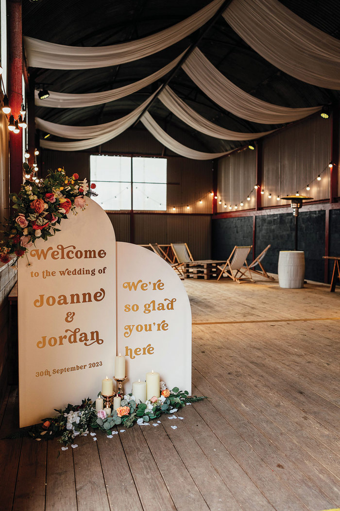 wedding welcome arched signage sits at the front of barn venue alongside an assortment of candles and flowers