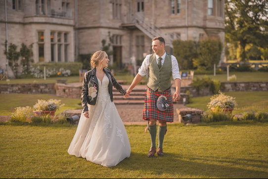 A bride wearing a white dress and a leather jacket holds hands with a groom wearing a red kilt walking across a lawn