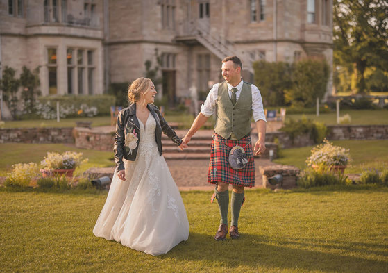 A bride wearing a white dress and a leather jacket holds hands with a groom wearing a red kilt walking across a lawn