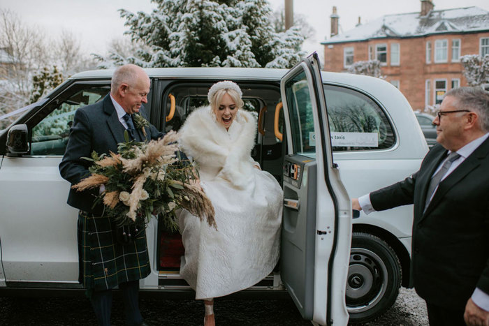 A bride stepping out of a silver taxi as a man in a suit holds the door open and a man in a kilt holds her bouquet