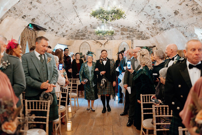 a groom wearing a kilt walking down an aisle on the arm of a woman wearing a teal dress as rows of guests watch on at Neidpath Castle