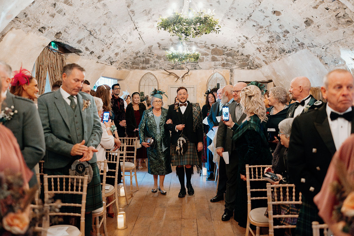 a groom wearing a kilt walking down an aisle on the arm of a woman wearing a teal dress as rows of guests watch on at Neidpath Castle