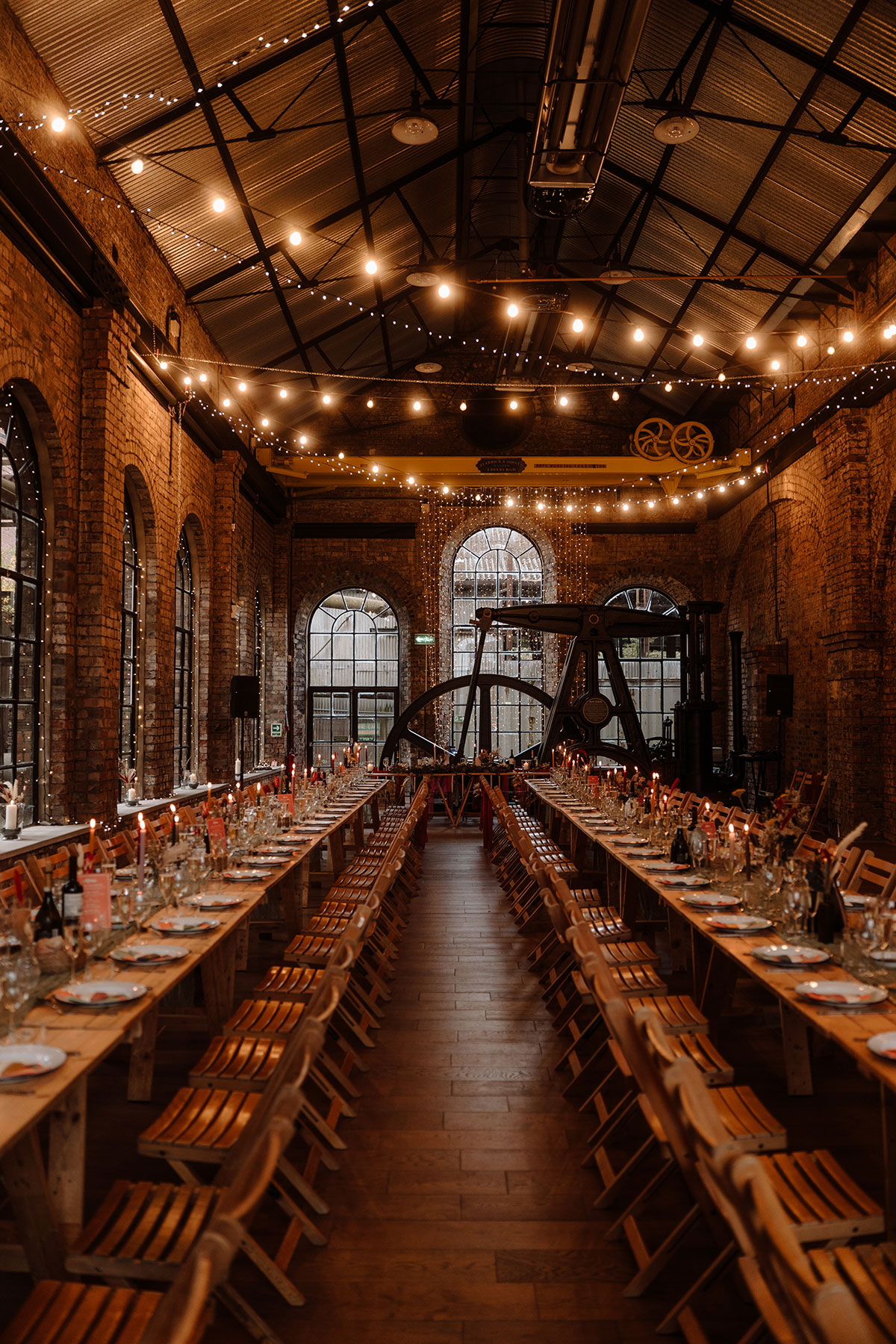 Long banquet tables styled for a wedding breakfast inside a restored industrial venue with exposed brickwork, fairy lights and historic machinery