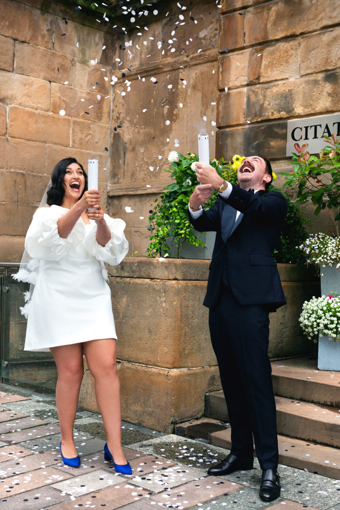 A bride in a short wedding dress and a groom in a dark suit set off confetti cannons filled with white confetti