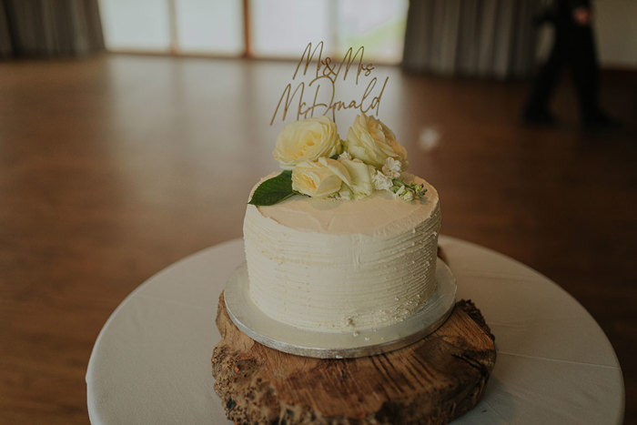 A Simple One Tier Buttercream Wedding Cake With Ivory Roses And Name Cake Topper Sitting On A Slice Of Wooden Log And Wooden Floor In Background