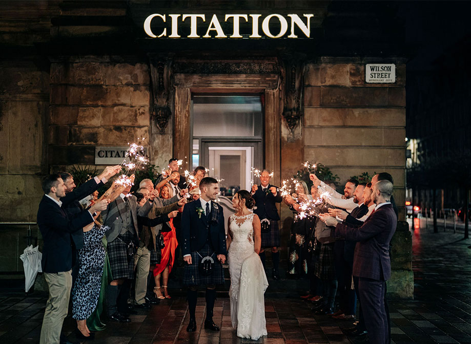 A bride and groom look at each other as they walk towards the camera out of a building as guests hold up sparklers