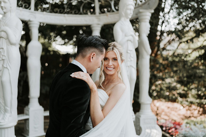 a bride and groom posing in garden at busby hotel under white ornamental pergola