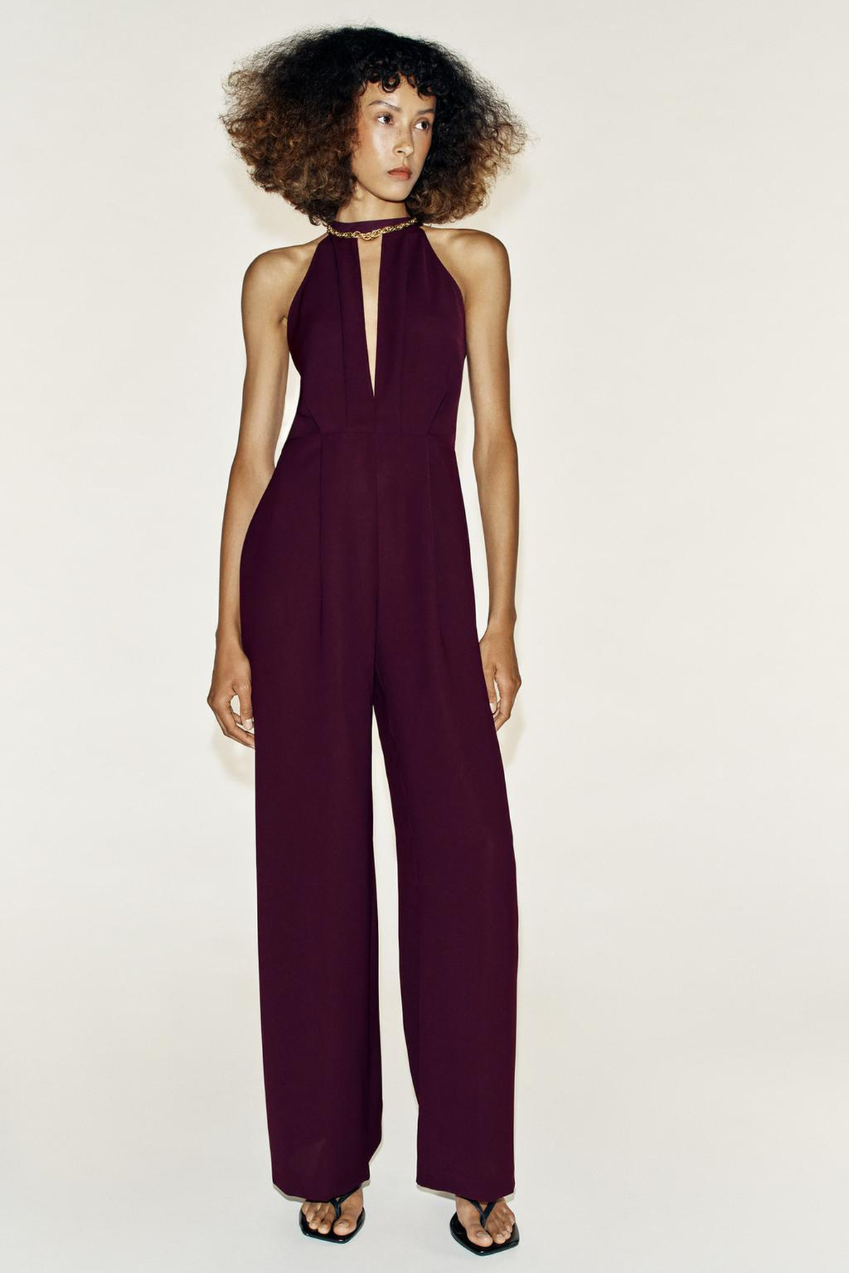 Model wearing a deep-plum halterneck jumpsuit with a plunging keyhole neckline, gold chain detail and wide-leg trousers.