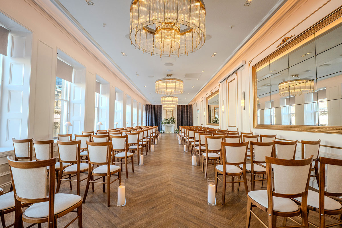 long room with dark wooden floor and brown and cream chairs lined in rows to create an aisle with candles lining each row and a fireplace and flowers at the very end