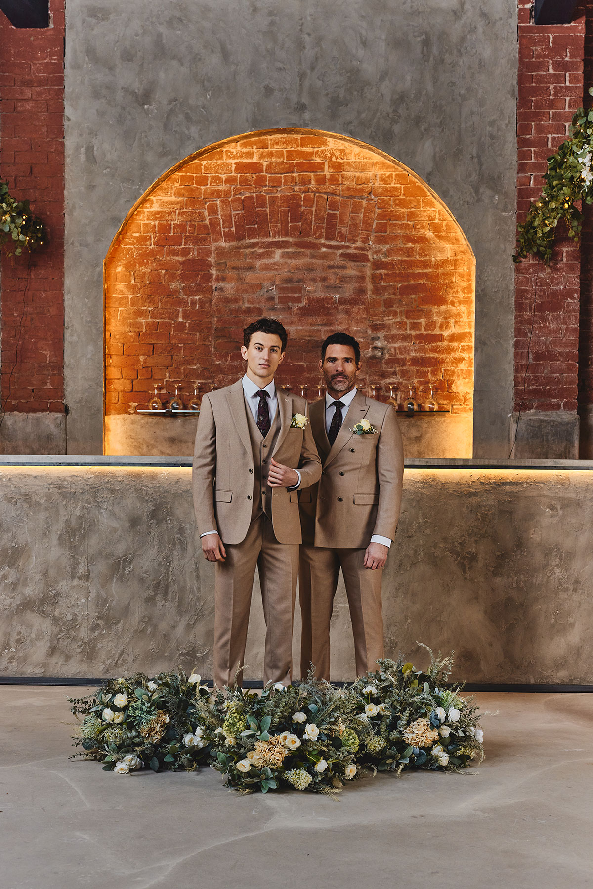 Two men standing in beige suits surrounded by flower decor