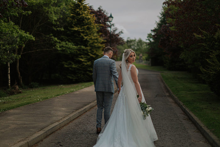 A Bride Looks Over Her Shoulder While Walking Along A Tree Lined Road Holding Hands With A Groom