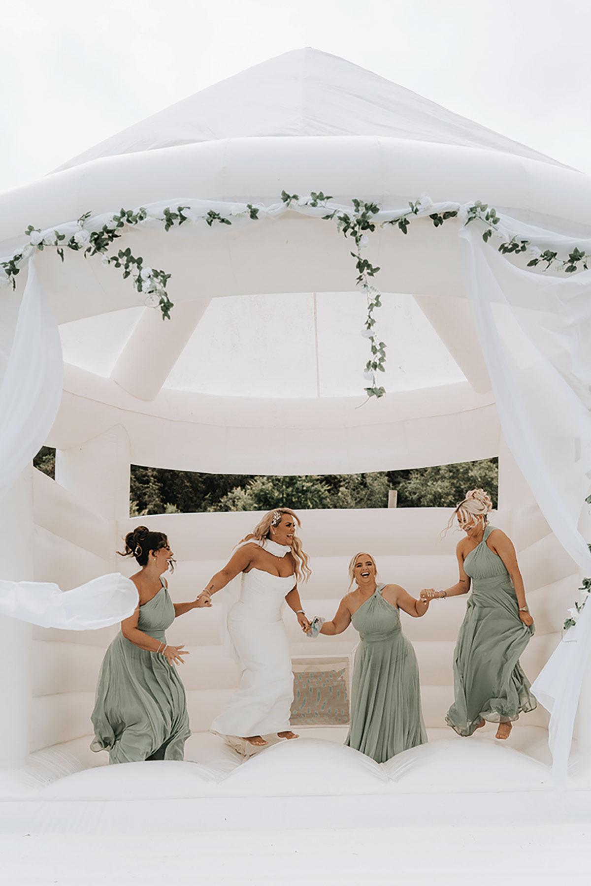 Bride and bridesmaids in sage-green dresses laughing and bouncing on a white wedding bouncy castle.