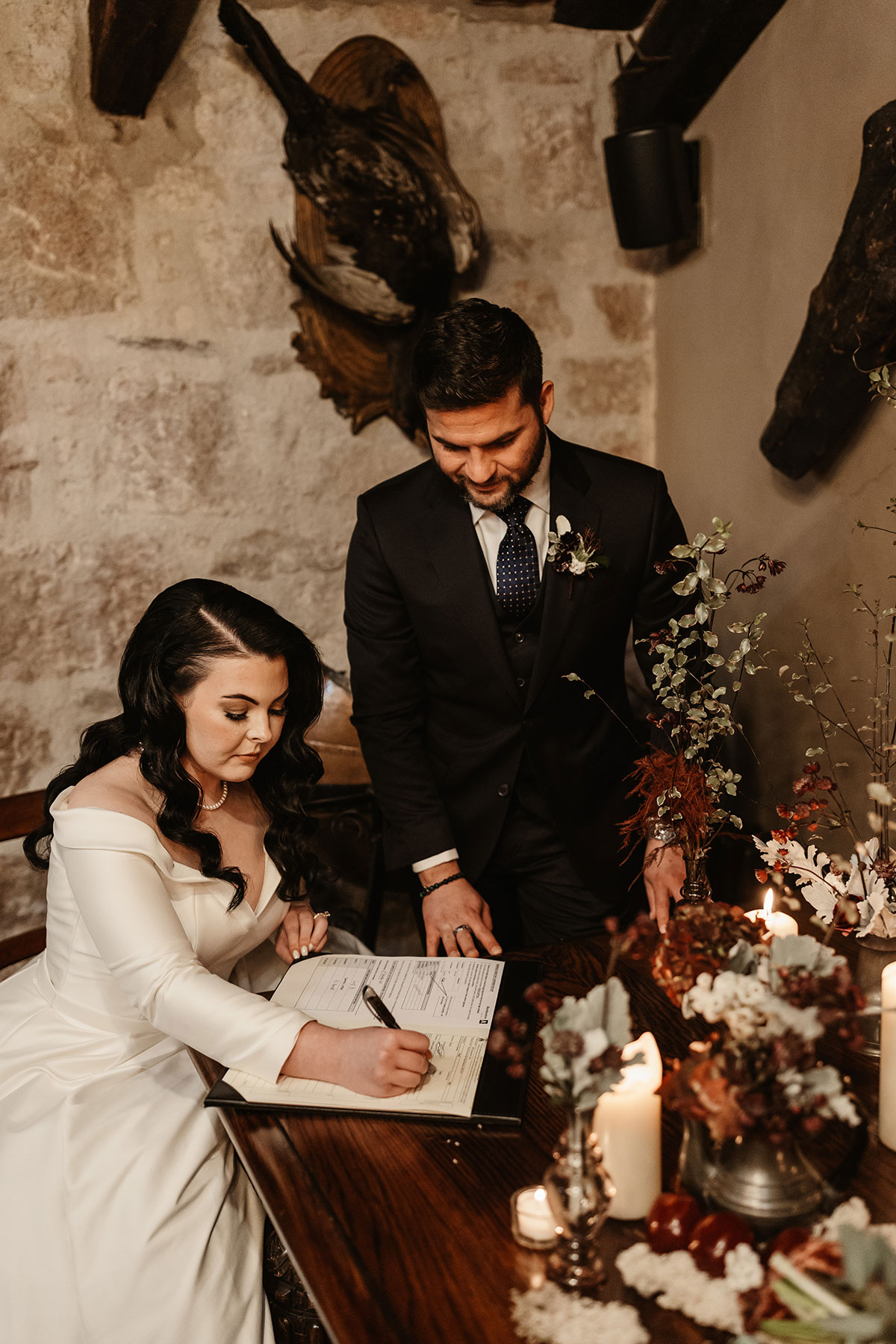 Bride signing the marriage register at a candlelit table as the groom stands beside her smiling