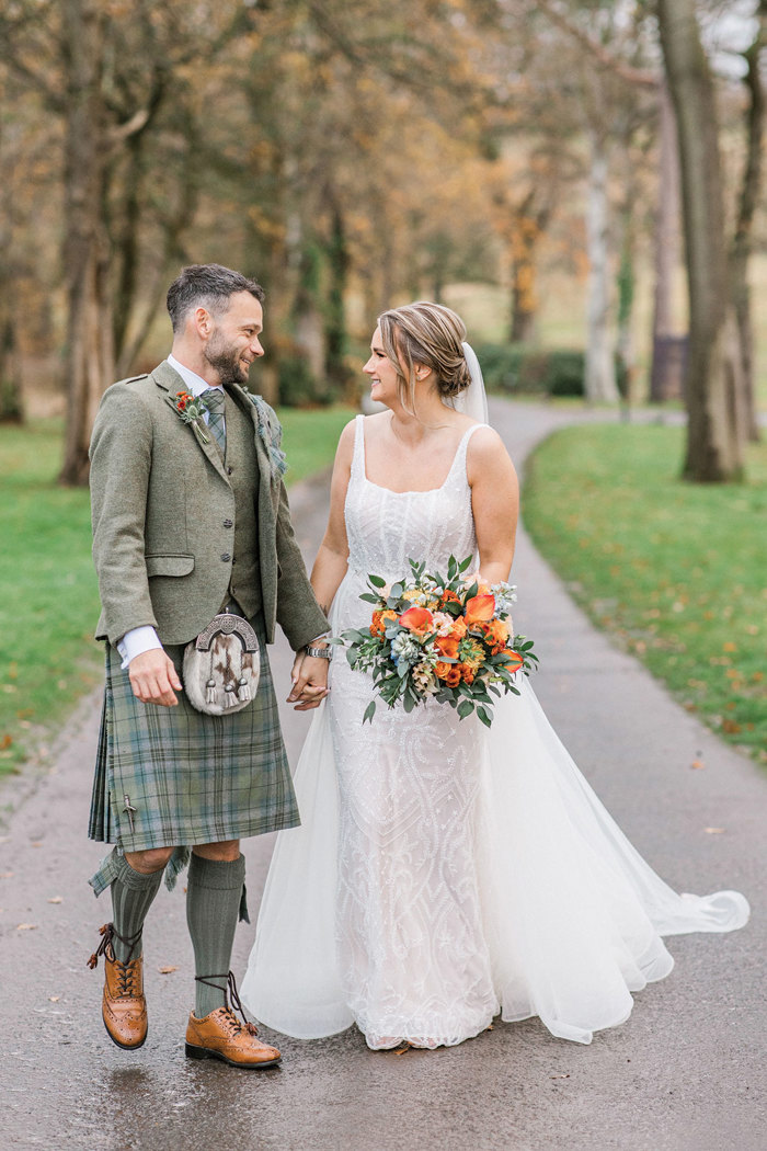 groom in green and blue tartan outfit and bride in beaded wedding dress with long tulle veil and trailing overskirt hold hands and look each other in the eyes outdoors on path