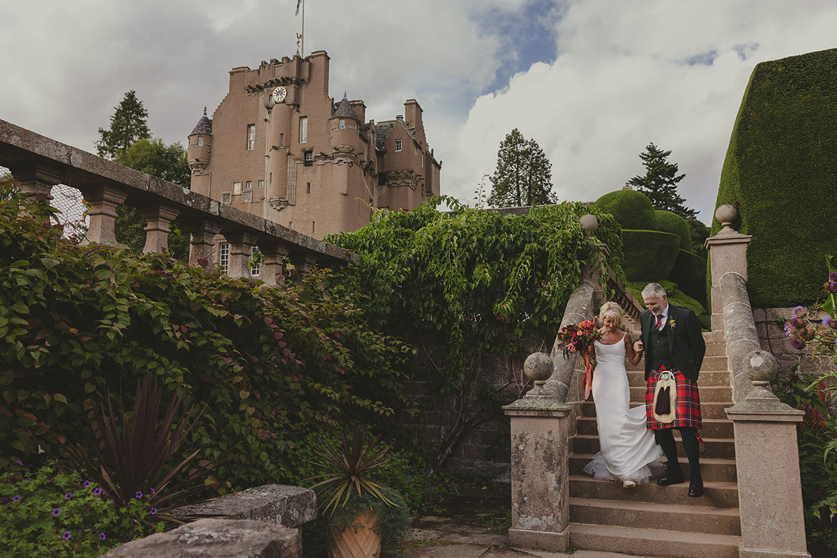 Bride and groom in kilt walking down stone garden steps surrounded by lush greenery, with castle visible behind them