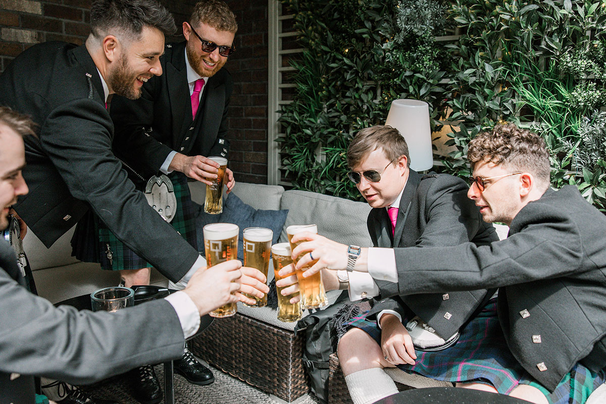Groomsmen in tartan kilts raising pints together before the wedding ceremony