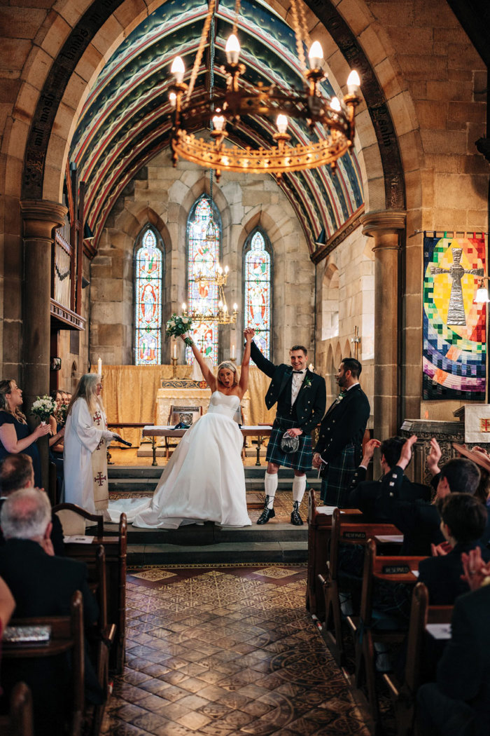 an elated bride and groom raising hands aloft in a church with stained glass windows