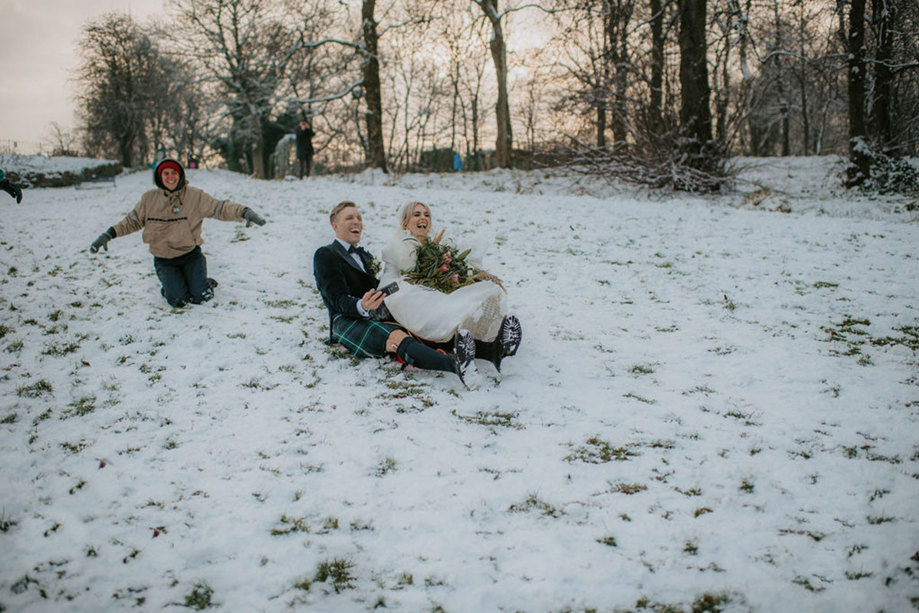 A bride wearing a white dress and white fur wrap and groom wearing a dark kilt sledging down a hill in the snow