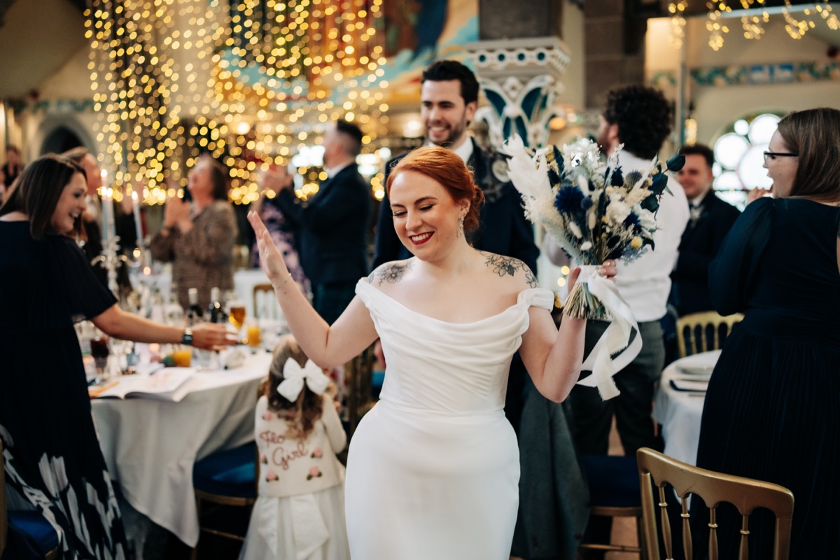 Bride Robyn enters her Òran Mór wedding reception carrying a blue-and-white bouquet as guests applaud beneath fairy lights.