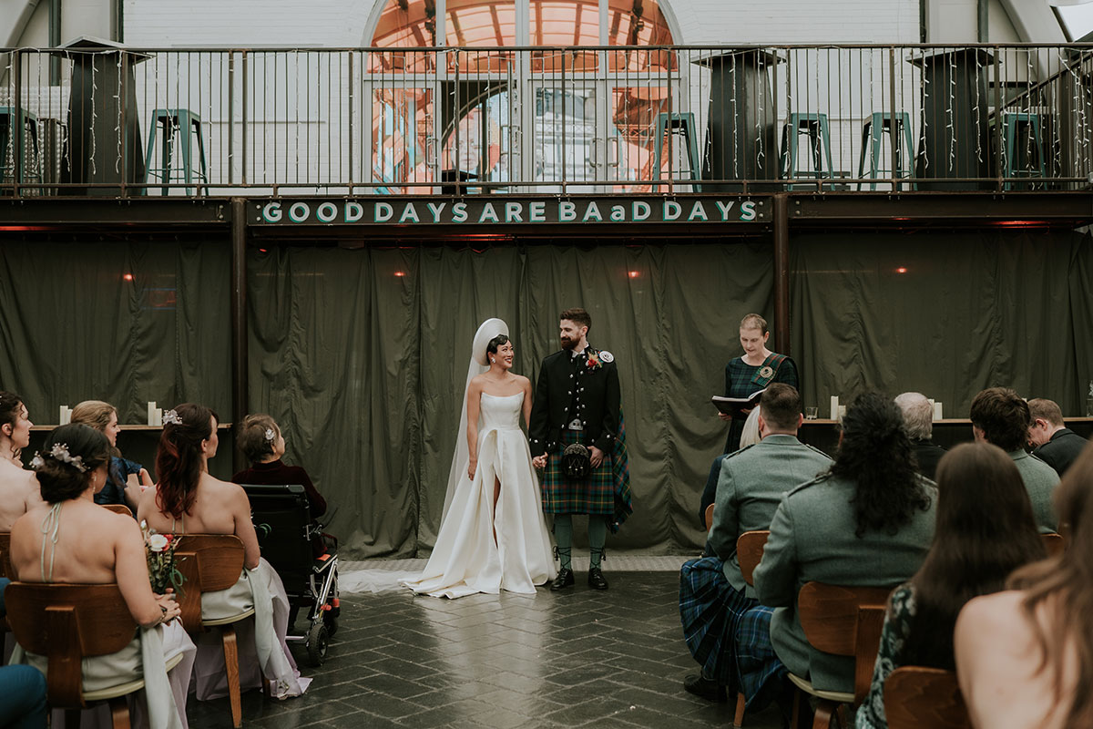 Wedding ceremony at Barras Art and Design Glasgow with bride, groom in kilt and guests seated indoors