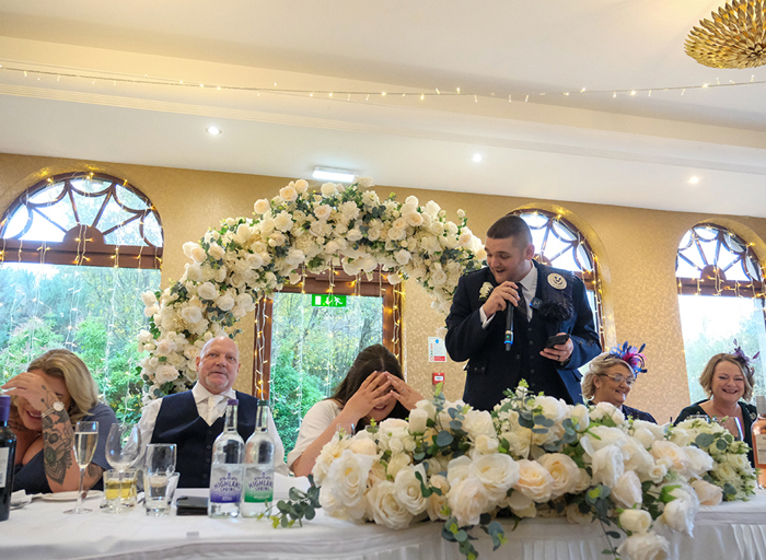 A groom making a speech holding a microphone and a phone while the bride and other people sitting at the table laugh