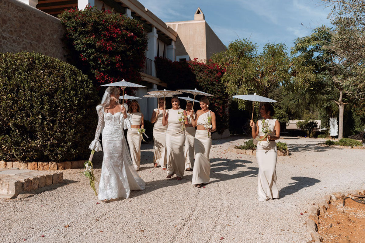 Bride and bridesmaids walking through Can Rafal Ibiza wedding venue holding white parasols before outdoor ceremony