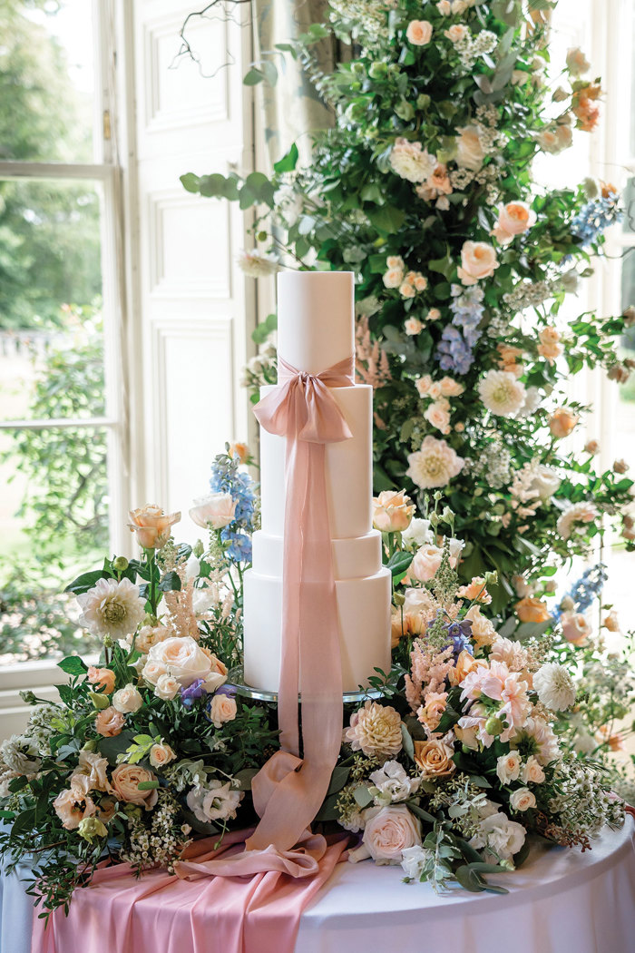 A white four tier wedding dress with a pink ribbon wrapped around the top tier and tied in a bow. Pastel flowers surround the base of the cake