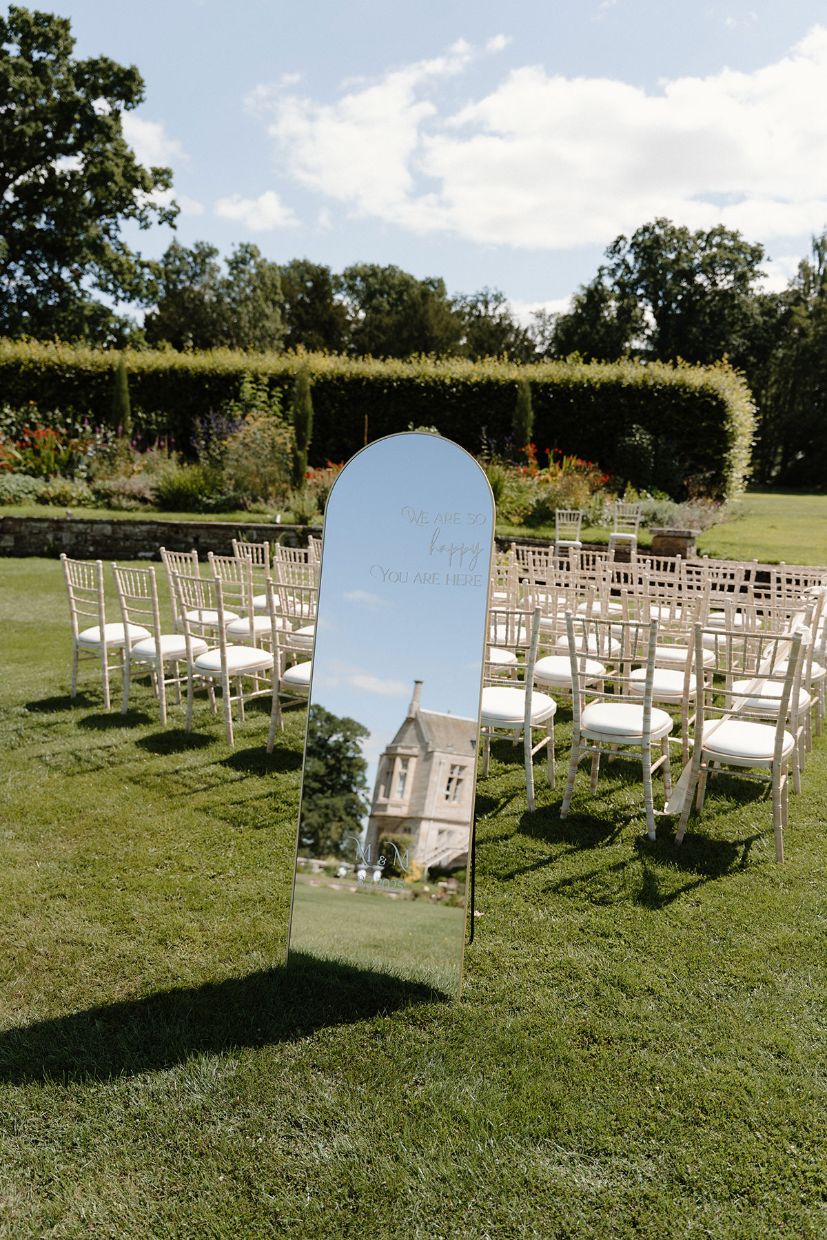 Outdoor wedding ceremony setup in a formal garden with mirrored welcome sign and elegant chiavari chairs on the lawn