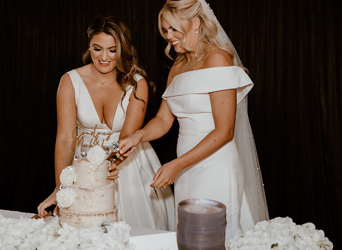 Two smiling brides cutting a wedding cake that is decorated with small disco balls, personalised cake topper and white flowers