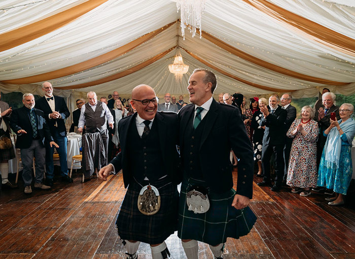 two grooms wearing kilts dancing in a marquee as guests stand on the side of the dance floor and clap