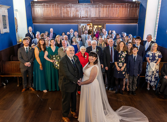 a smiling bride and groom standing in front of a group of wedding guests in a church hall 