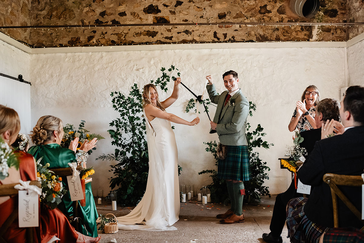 Couple tying the knot during the handfasting ceremony, smiling as guests applaud inside a rustic stone barn