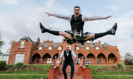 a groom wearing a kilt jumping in front of another groom in front of the exterior of Boclair House