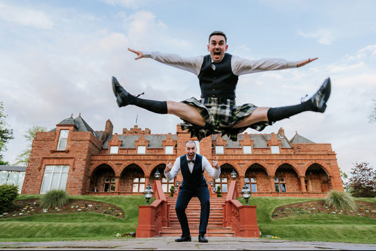 a groom wearing a kilt jumping in front of another groom in front of the exterior of Boclair House