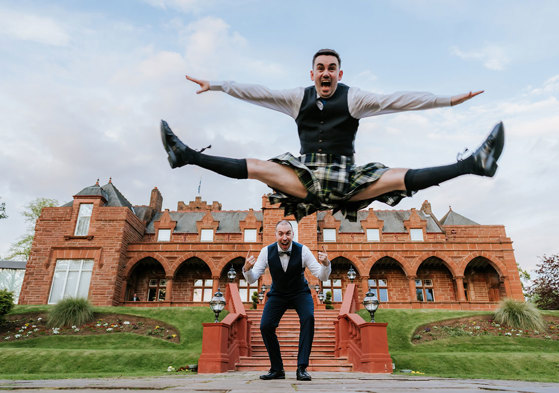 a groom wearing a kilt jumping in front of another groom in front of the exterior of Boclair House