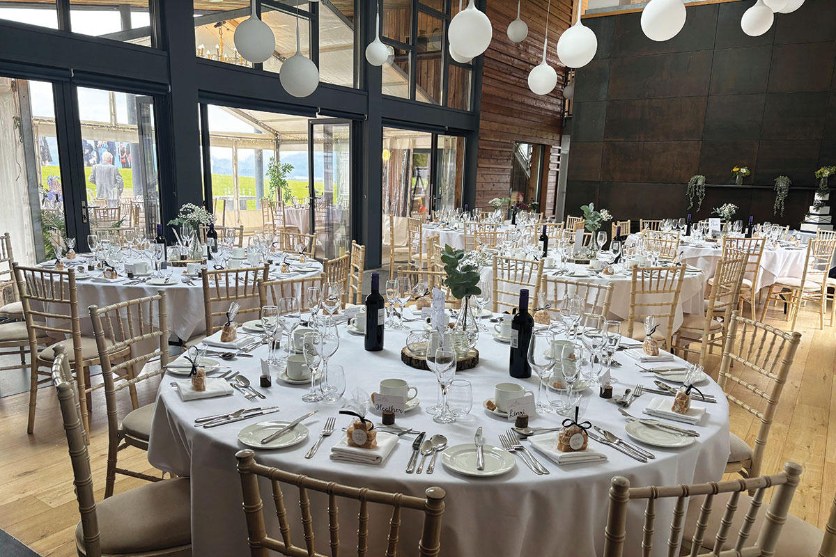 a room is filled with round tables, all set with white linens for dining, chairs set around them and large bulb statement light features hanging from the ceiling