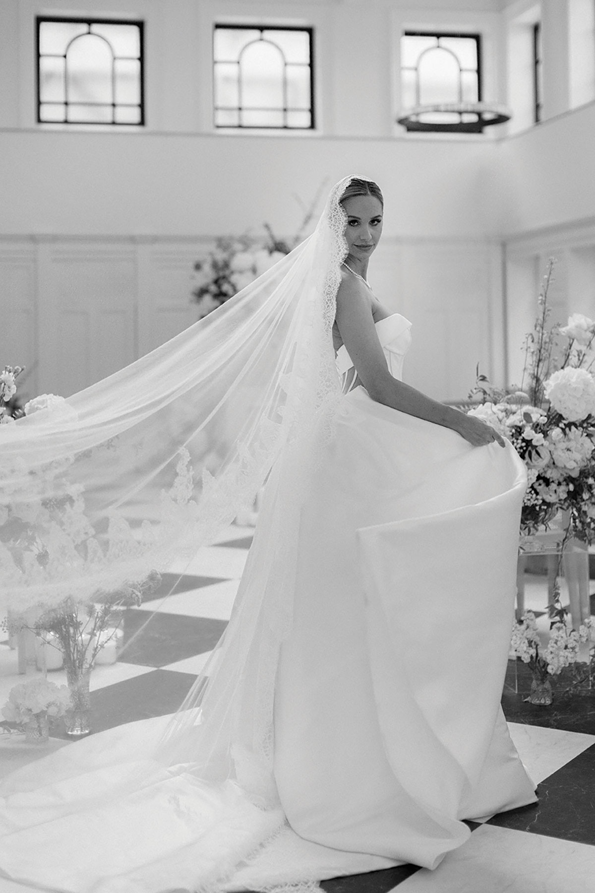 Black-and-white photo of bride in strapless gown with lace veil flowing down aisle