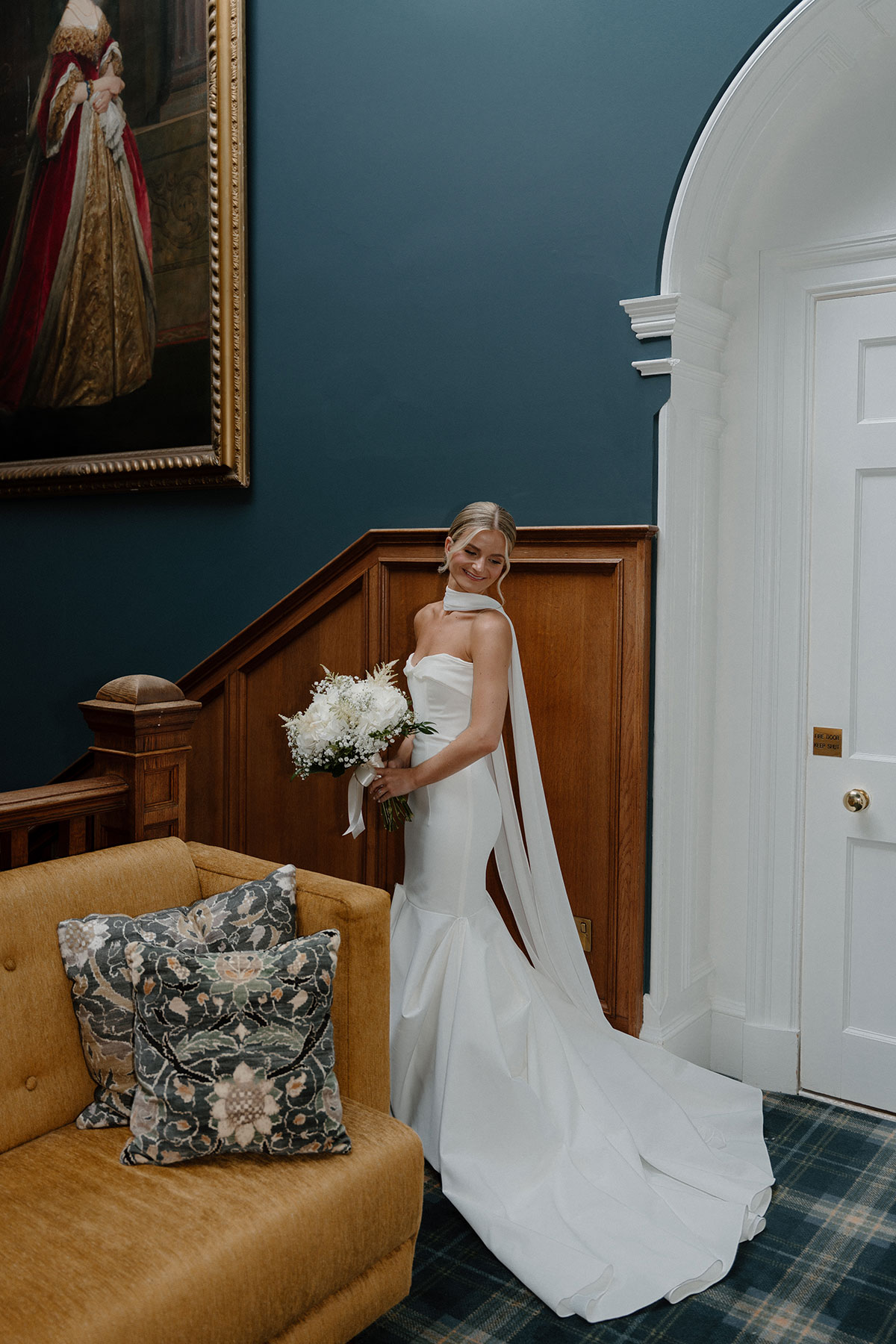 Bride in a fitted satin wedding dress holding a white bouquet inside a stylish country house wedding venue with dark blue walls and wood panelling