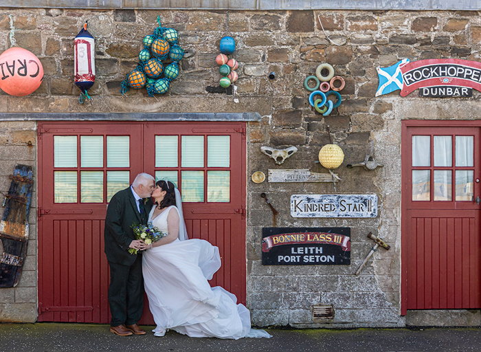 a bride and groom kissing in front of the red doors of a boat shed decorated with sea paraphernalia