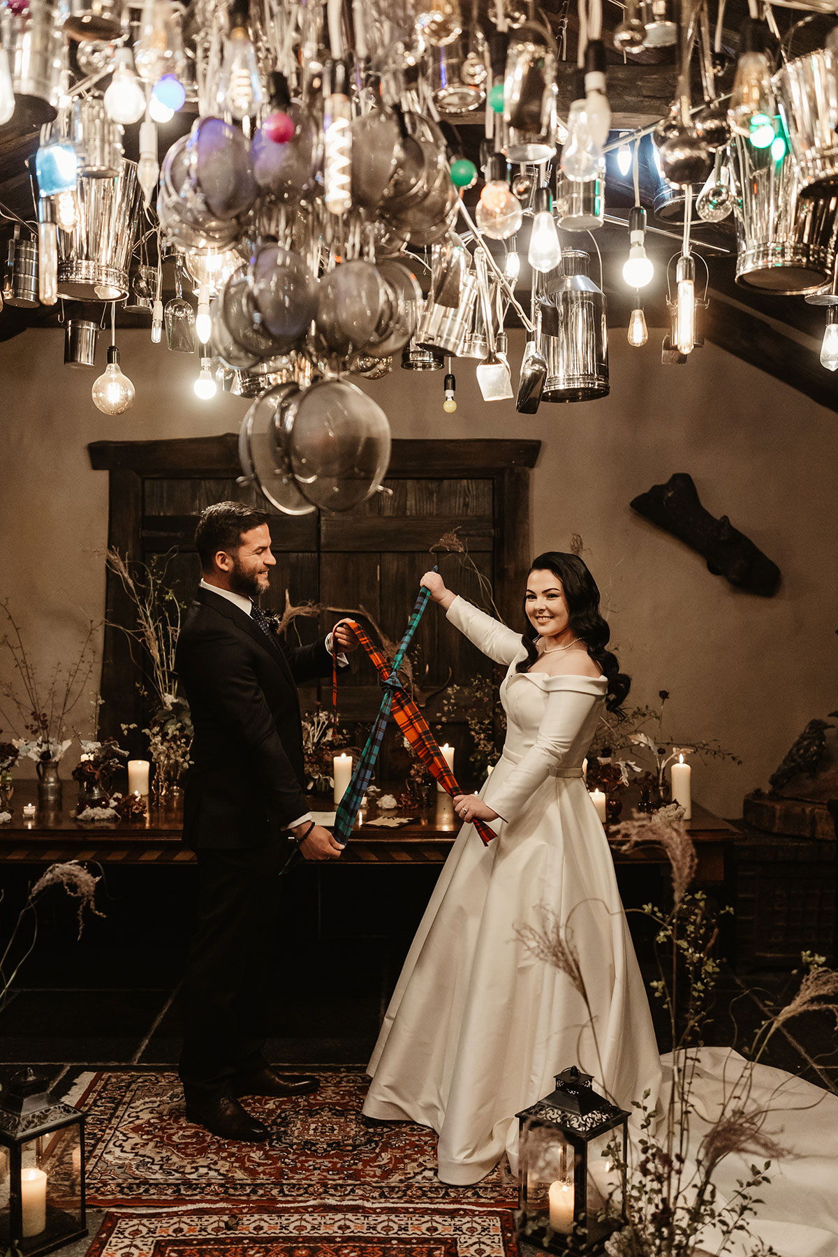 Bride and groom lifting tartan ribbons together in the ceremony room, surrounded by candles and eclectic lighting above